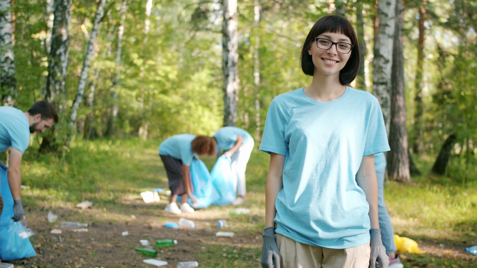 A volunteer in gloves at a forest cleanup, with others bagging trash behind her